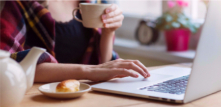 A young woman uses her laptop at home while she enjoys a coffee and a croissant. 
